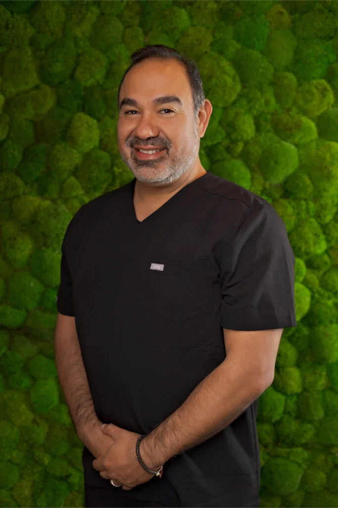 Dr. Gallegos in black medical scrubs smiling against a vibrant green moss backdrop, representing Thrive Medicine Clinic's focus on personalized wellness and testosterone replacement therapy.