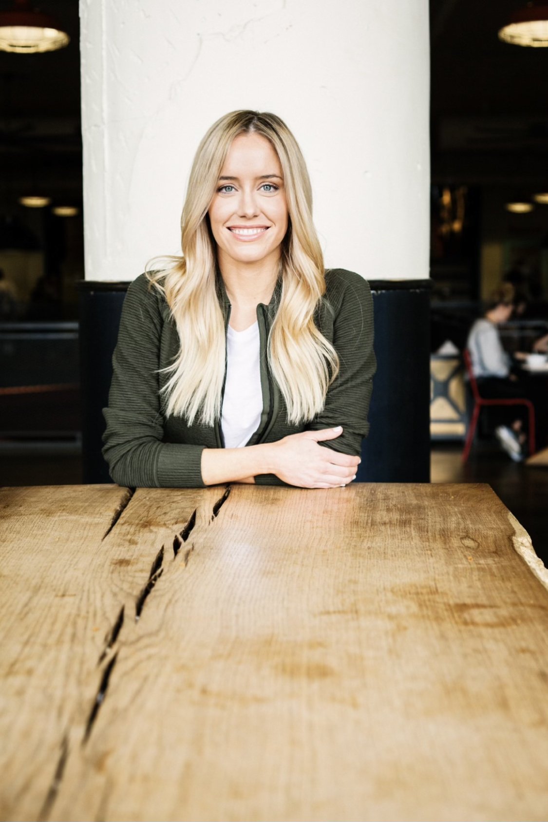 Smiling woman with long blonde hair, wearing a green jacket, sitting at a wooden table, representing the welcoming environment of Thrive Medicine Clinic and its focus on personalized patient care.