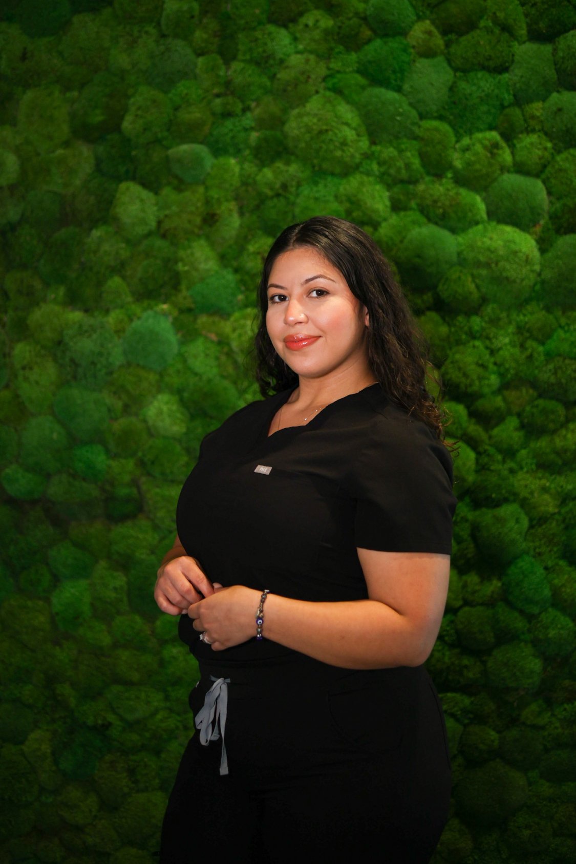 Leslie, a compassionate healthcare professional at Thrive Medicine Clinic, posing in front of a vibrant green moss wall, embodying the clinic's commitment to personalized care and wellness.