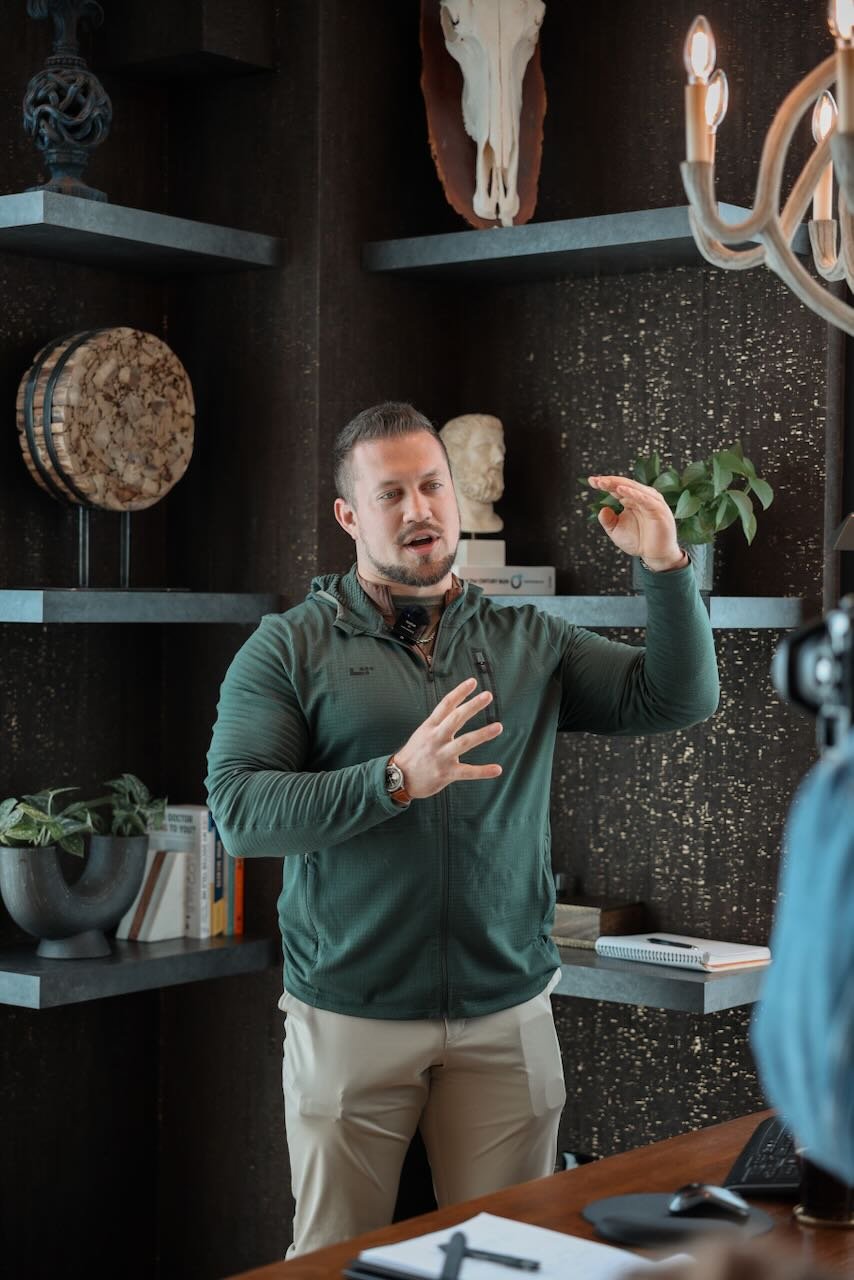 Man in green jacket explaining non-surgical penile enhancement options in a clinic setting, with books and plants in the background.