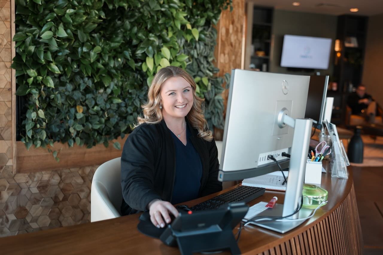Smiling receptionist at Thrive Medicine Clinic's front desk, surrounded by greenery, with computer and office supplies, representing patient care and wellness consultation.