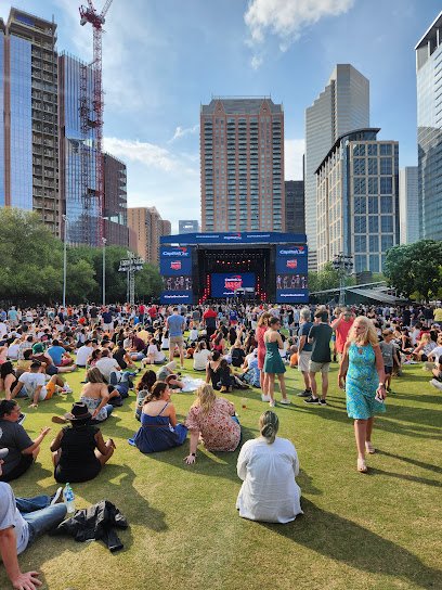 Crowd gathered on grassy area in urban park for outdoor event, large stage with screens in background, surrounded by modern buildings.