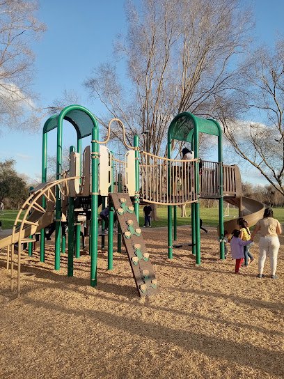 Playground structure with climbing features, slide, and children playing in a park setting, surrounded by trees and open space.